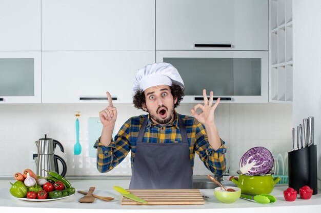Front view of scared male chef with fresh vegetables and cooking with kitchen tools and making eyeglasses gesture pointing up in the white kitchen