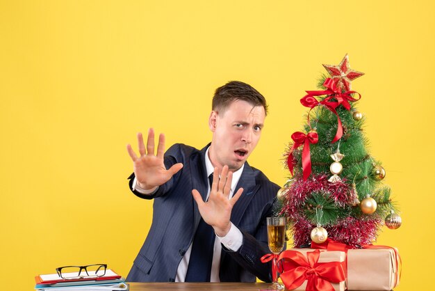 Front view of scared man rejecting something sitting at the table near xmas tree and presents on yellow