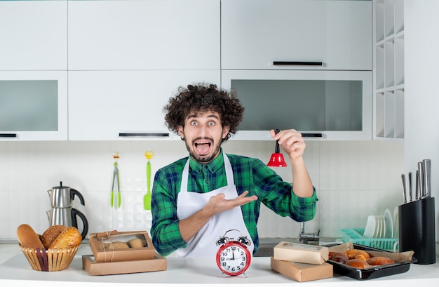 Front view of scared young man standing behind the table various pastries on it and showing red ring bell in the white kitchen