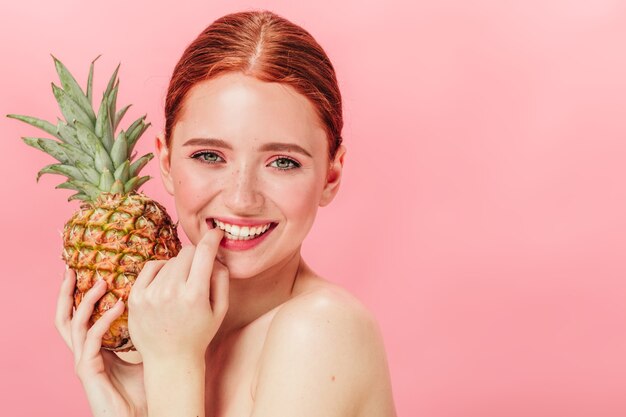 パイナップルを保持している官能的なヨーロッパの女性の正面図。カメラで笑っている果物と幸せな女の子のスタジオショット。