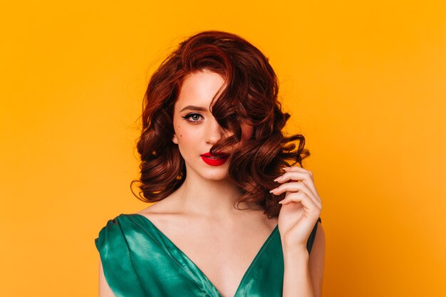 Front view of sensual woman with ginger wavy hair. Studio shot of elegant girl in green dress isolated on yellow space.