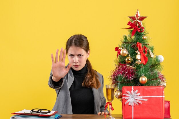 Front view serious girl sitting at the table making stop gesture near xmas tree and gifts cocktail