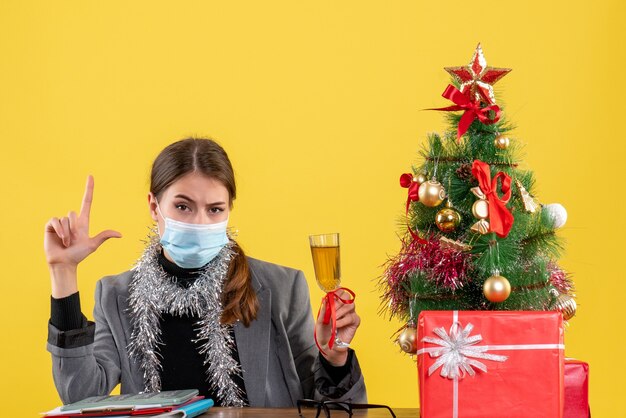 크리스마스 트리와 선물 칵테일을 홀 짝 테이블에 앉아 의료 마스크와 전면보기 심각한 소녀