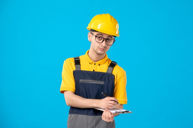 Front view of serious male builder in uniform taking notes on blue 