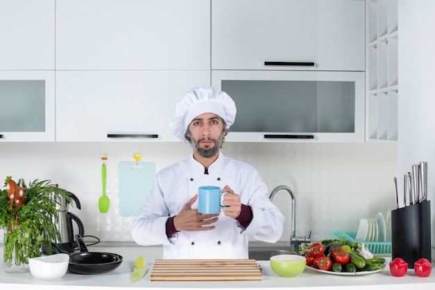 キッチンテーブルの後ろに立っているカップを保持しているコック帽子の正面図真面目な男性シェフ