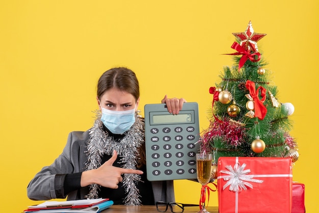 손가락 계산기 크리스마스 트리와 선물 칵테일 지적 테이블에 앉아 의료 마스크 전면보기 심각한 어린 소녀