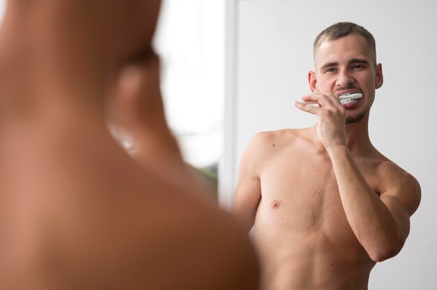 Вид спереди человека без рубашки, чистящего зубы в зеркале