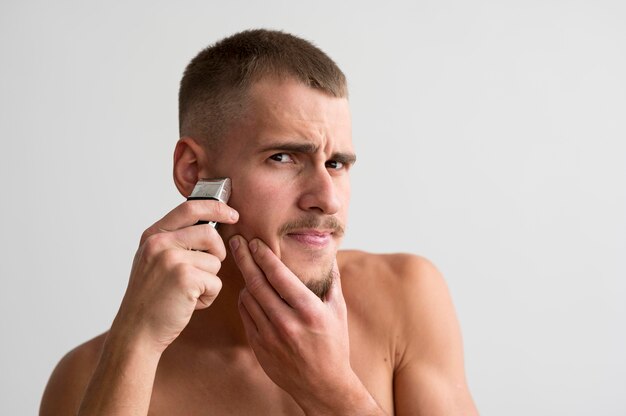 Front view of shirtless man using electric shaver for his beard