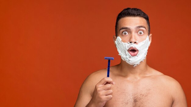 Front view of shirtless man with shaving foam on his face holding razor