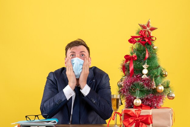 Front view of shocked business man looking up sitting at the table near xmas tree and presents on yellow