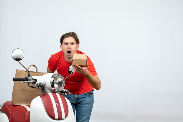 Front view of shocked delivery guy in red uniform standing near scooter giving order on white background