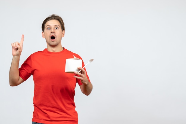 Вид спереди потрясенного эмоционального молодого парня в красной блузке, держащего бумажную коробку и указывающего вверх на белом фоне