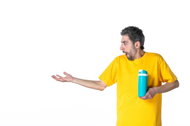 Front view of shocked emotional young male in yellow shirt and showing blue thermos pointing something on the right side on white background