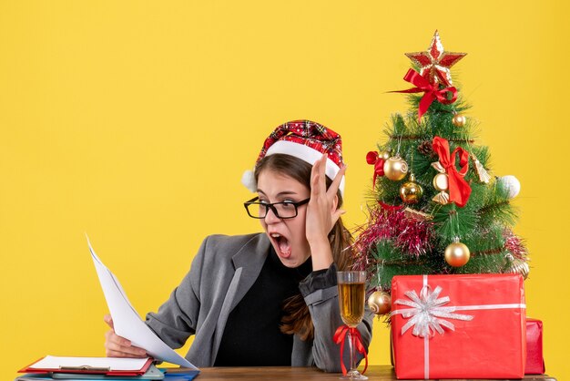 Вид спереди потрясенная девушка в рождественской шляпе, сидящая за столом, глядя на документы, рождественскую елку и коктейль с подарками