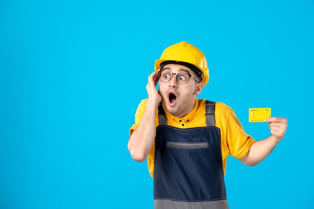 Front view of shocked male builder in uniform and helmet with credit card on a blue 