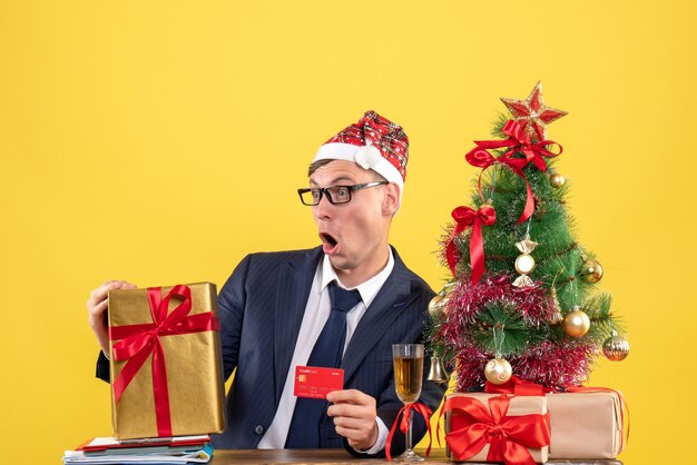 Вид спереди потрясенного человека, держащего карточку, сидящего за столом возле рождественской елки и подарков на желтом