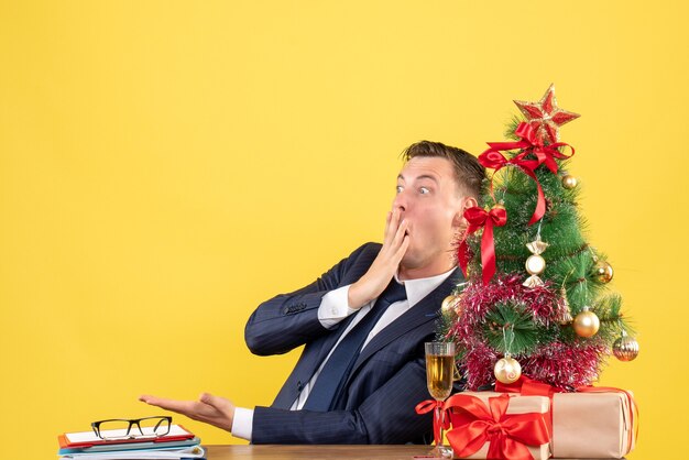 クリスマスツリーの近くのテーブルに座って、黄色で提示するショックを受けた男の正面図。