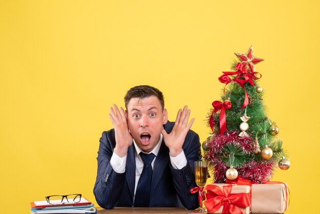 クリスマスツリーの近くのテーブルに座って、黄色で提示するショックを受けた男の正面図。