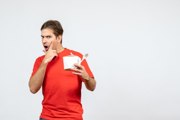 Front view of shocked young guy in red blouse holding paper box and spoon on white background