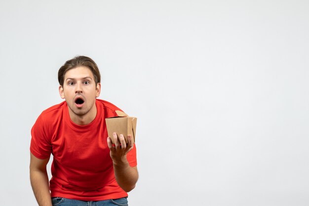 Front view of shocked young guy in red blouse holding small box on white background