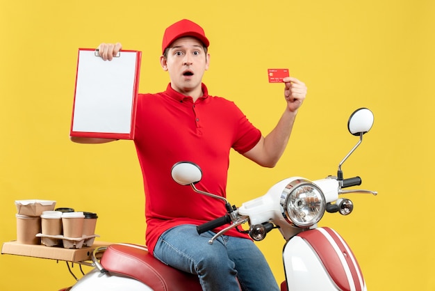 赤いブラウスと黄色の背景にドキュメントと銀行カードを保持している注文を配信銀行カードを身に着けているショックを受けた若い男の正面図