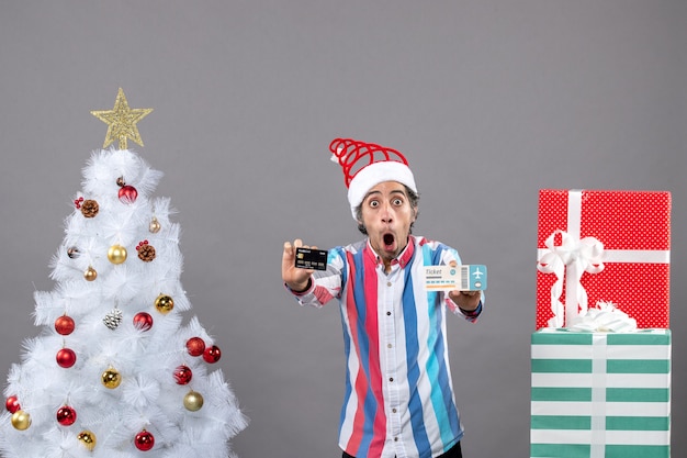 Front view shocked young man holding card and travel ticket around xmas tree and presents