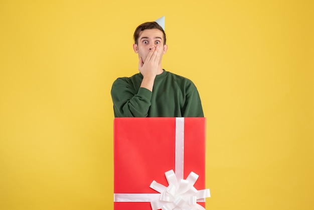 Free photo front view shocked young man with party cap standing behind big giftbox on yellow