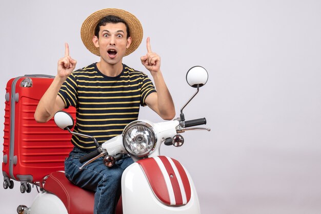 Front view of shocked young man with straw hat on moped pointing at ceiling