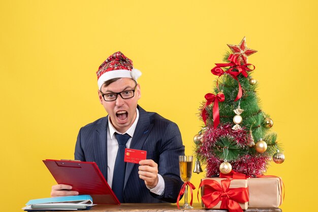 Front view of shouted business man holding clipboard sitting at the table near xmas tree and presents on yellow.