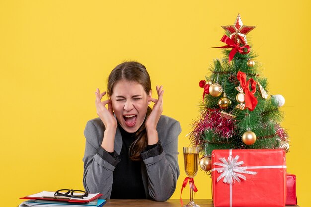 Front view shouted girl sitting at the desk covering ears with her fingers xmas tree and gifts cocktail