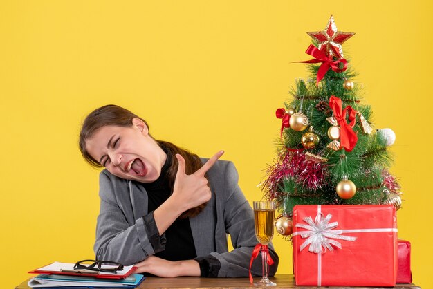 Front view shouted girl sitting at the desk pointing with finger xmas tree and gifts cocktail