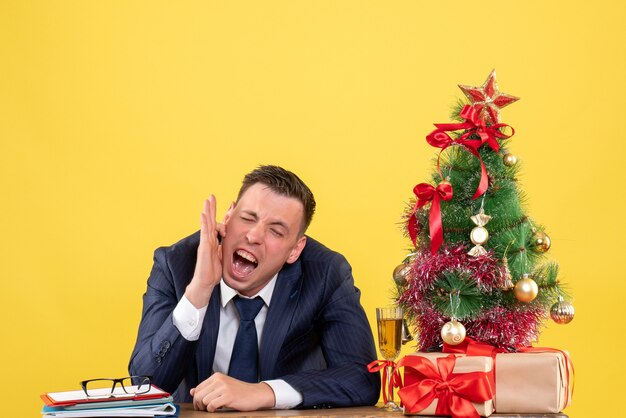 クリスマスツリーと黄色の贈り物の近くのテーブルに座って彼の耳を保持している叫んだ男の正面図。