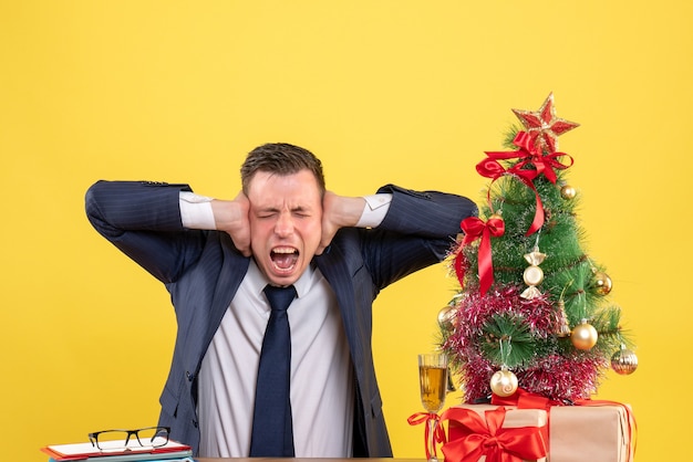 Front view of shouted man holding his ears sitting at the table near xmas tree and gifts on yellow.