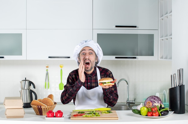 Front view of shouted man holding up burger standing behind kitchen table