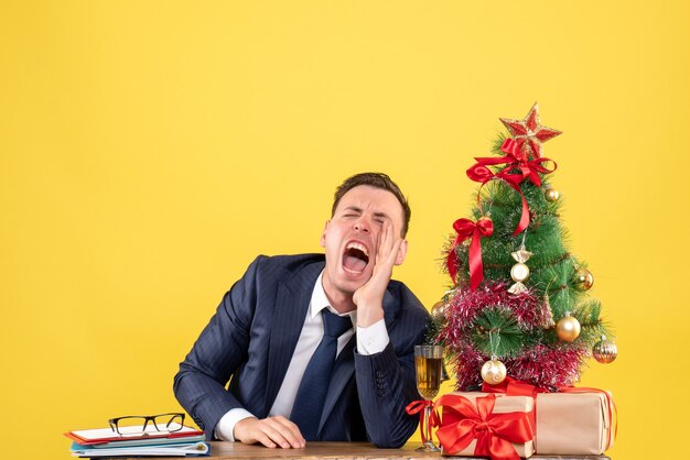クリスマスツリーと黄色の贈り物の近くのテーブルに座って叫んでいる男の正面図
