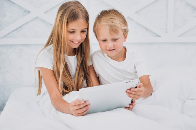 Front view siblings staying in bed while playing on a tablet