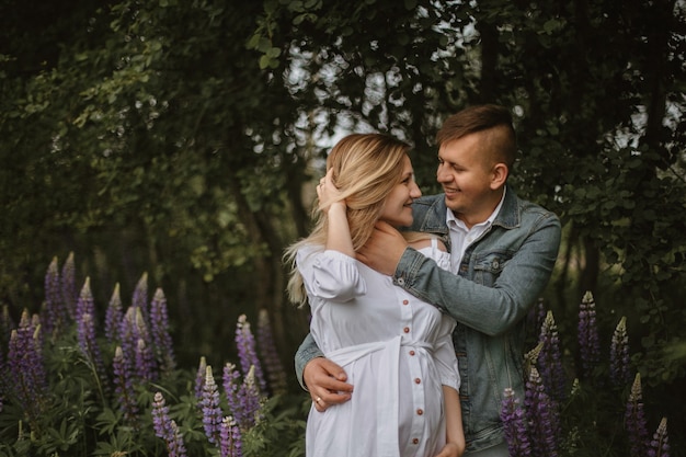 Front view of sincere smiled happy couple in green park with violet lupine, expecting baby