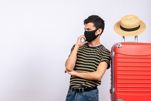 Front view skeptical young tourist with black mask standing near red suitcase