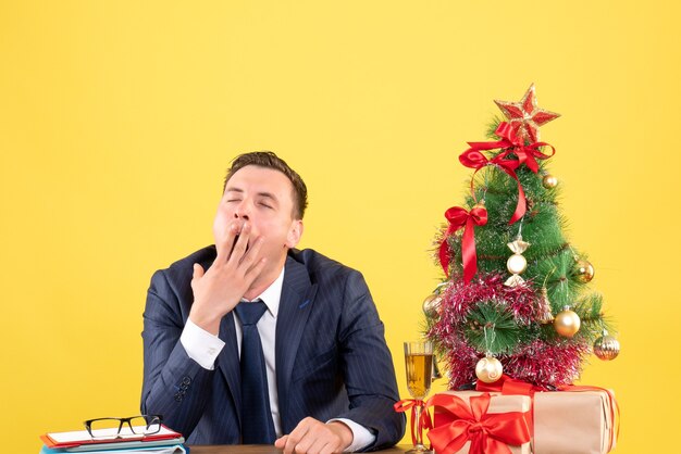 Front view of sleepy man yawning sitting at the table near xmas tree and gifts on yellow