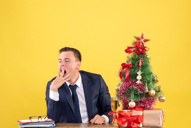 Front view of sleepy man yawning while sitting at the table near xmas tree and gifts on yellow