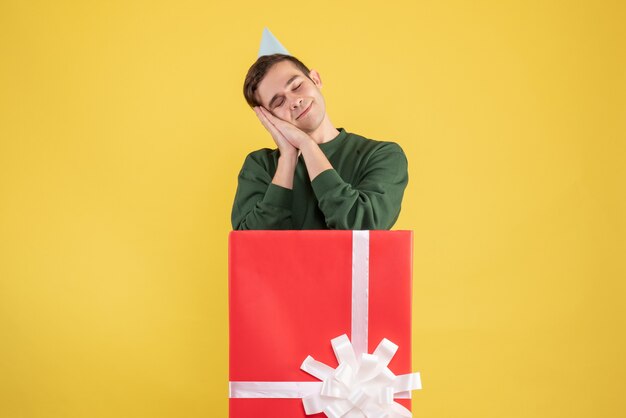 Front view sleepy young man with party cap standing behind big giftbox on yellow 