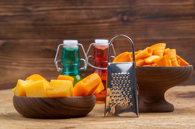 Front view slices of cheese and chips in bowls grater red and green bottles on brown isolated surface
