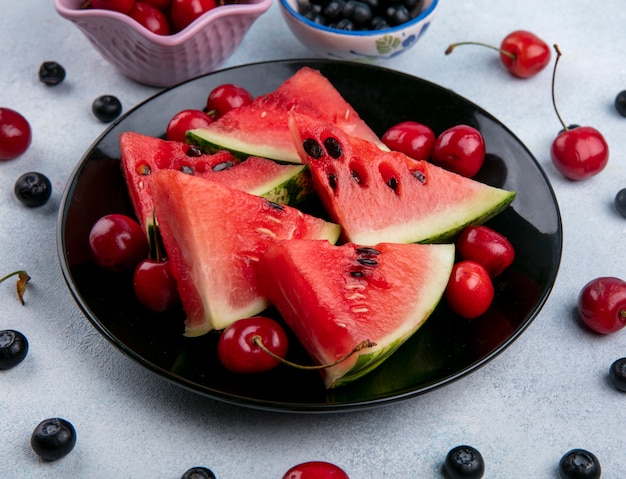 Front view slices of watermelon on a black plate with blueberries and cherries