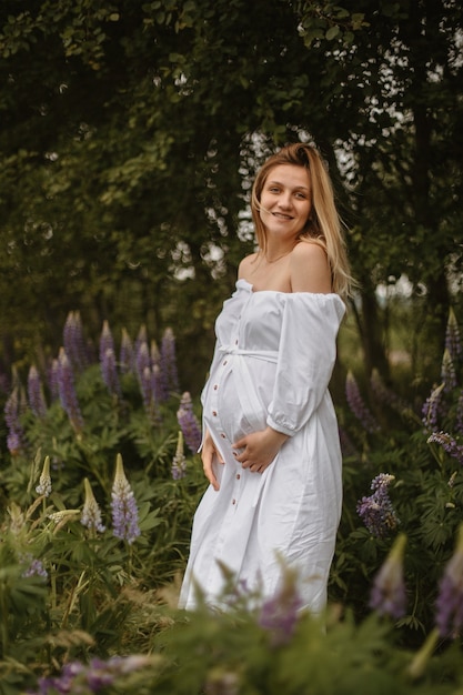 Front view of smiled blonde caucasian girl expecting first child looking straight, touching belly surrounded with lupines