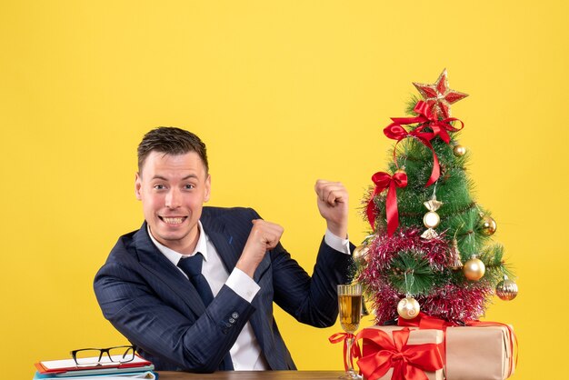 クリスマスツリーと黄色の贈り物の近くのテーブルに座って後ろを指している笑顔の男の指の正面図