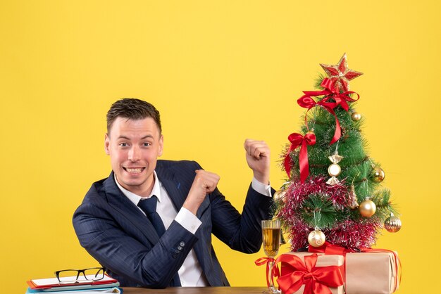 Front view of smiled man finger pointing back sitting at the table near xmas tree and gifts on yellow