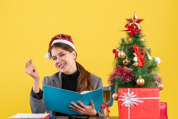 正面図は、テーブルに座っているクリスマス帽子とクリスマスツリーとギフトカクテルと笑顔の若い女の子