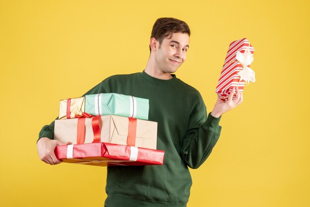 正面図は黄色の上に立っている新年の贈り物を保持している緑のセーターで若い男を微笑んだ