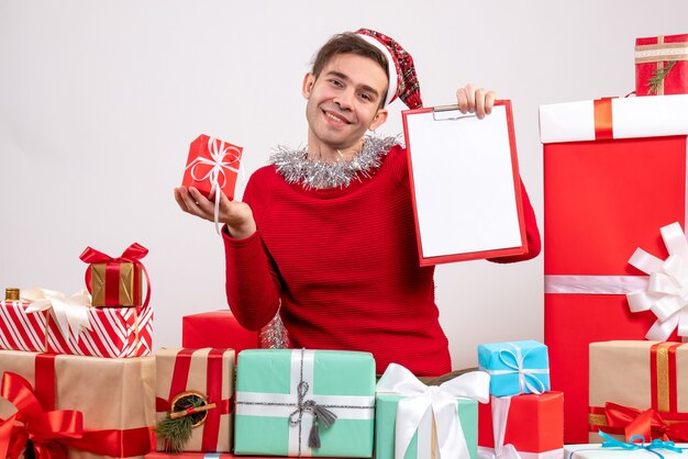 Вид спереди улыбающегося молодого человека в новогодней шапке, сидящего вокруг рождественских подарков