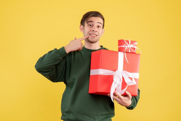 Вид спереди улыбающегося молодого человека с рождественским подарком, указывающего на его улыбку, стоящую на желтом